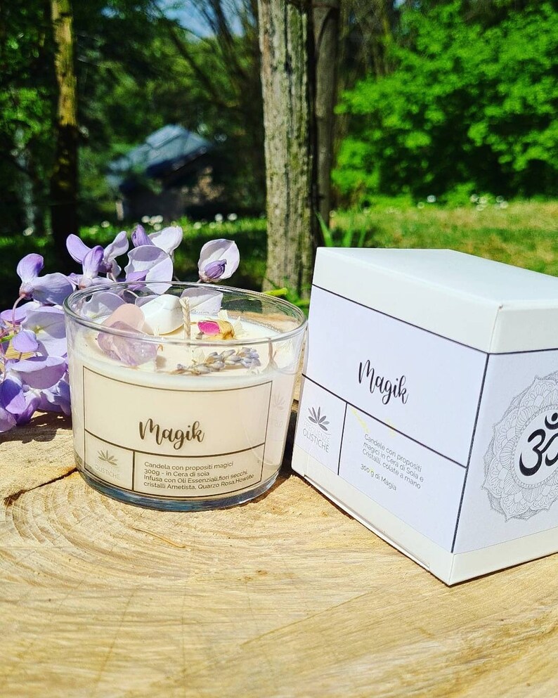 May include: A white candle jar with the word "Magik" printed on the label. The candle is infused with crystals and essential oils. The jar is sitting on a wooden surface with a white box next to it. The box also has the word "Magik" printed on it.