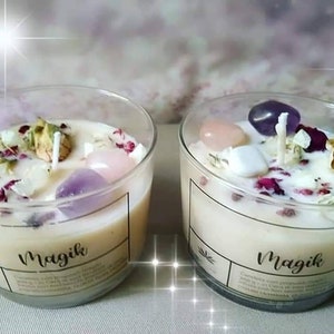 May include: Two clear glass candle jars with white wax and embedded crystals, dried flowers, and a wick. The jars have a black label with the word "Magik" printed on them.