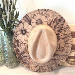 May include: A beige fedora hat with a floral and feather design etched into the brim. The hat features a soft, suede-like texture and a small tassel. The hat is displayed on a light-colored surface.