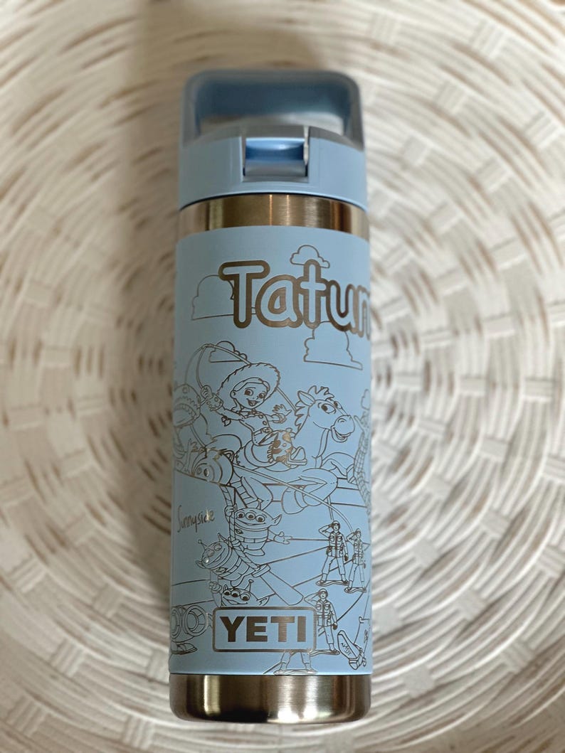 May include: A light blue Yeti water bottle with a stainless steel lid and a white outline of Toy Story characters on the side. The bottle has the text "Tatum" and "YETI" on it.