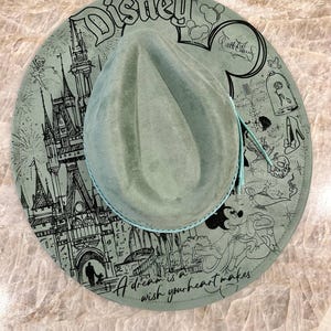May include: A sage green fedora-style hat with a wide brim featuring a Disney theme. The brim is decorated with black line art of Disney characters and landmarks, including a castle and Mickey Mouse. The text "Disney" and "A dream is a wish your heart makes" are also visible.
