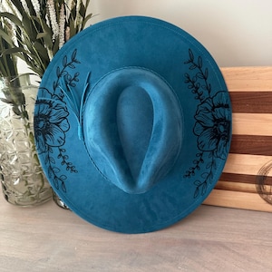 May include: A teal fedora-style hat with a wide brim. The hat features black floral and leaf designs on the brim. A small feather is attached to the side. The hat is displayed on a wooden surface.