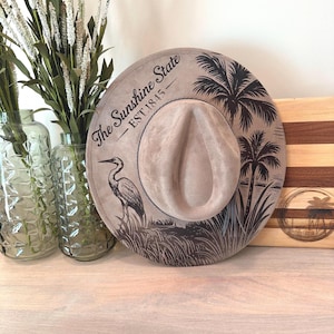 May include: A tan fedora hat with a wide brim. The hat features a heron and palm tree design with the text "The Sunshine State - EST 1845". The hat is displayed on a wooden surface.