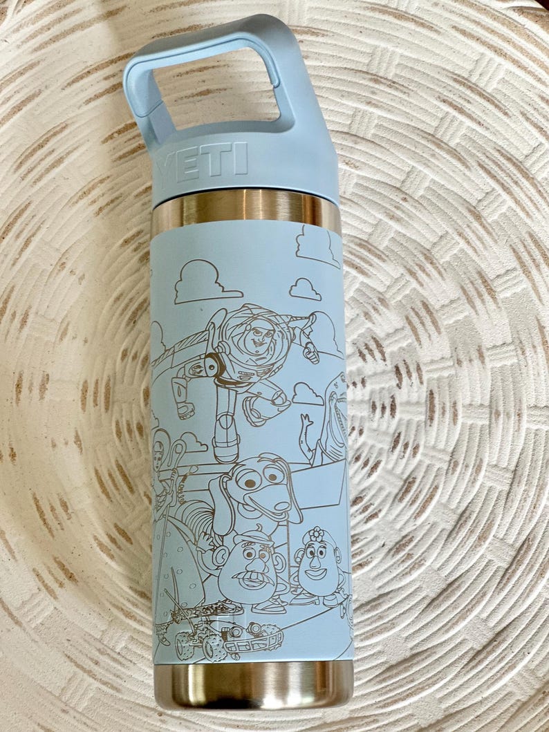 May include: A light blue Yeti water bottle with a stainless steel body and a black and white outline of Toy Story characters on the side. The bottle has a black handle and the word "YETI" is printed on the top.