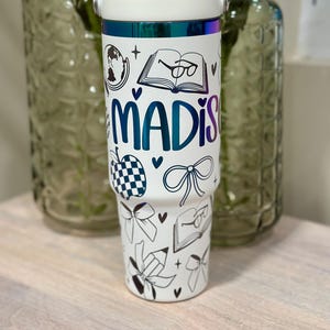 May include: White insulated tumbler with a handle, featuring the name "MADIS" in teal and black line art illustrations of books, bows, and an apple. The top rim has an iridescent finish.