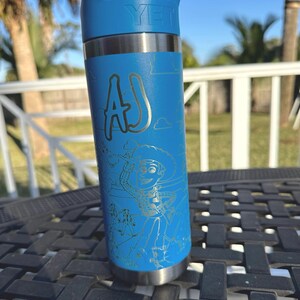 May include: A blue Yeti water bottle with a silver lid and a silver base. The bottle has a white inscription that says "YETI" and a silver inscription that says "AJ". The bottle also has a silver engraved image of Woody from Toy Story.