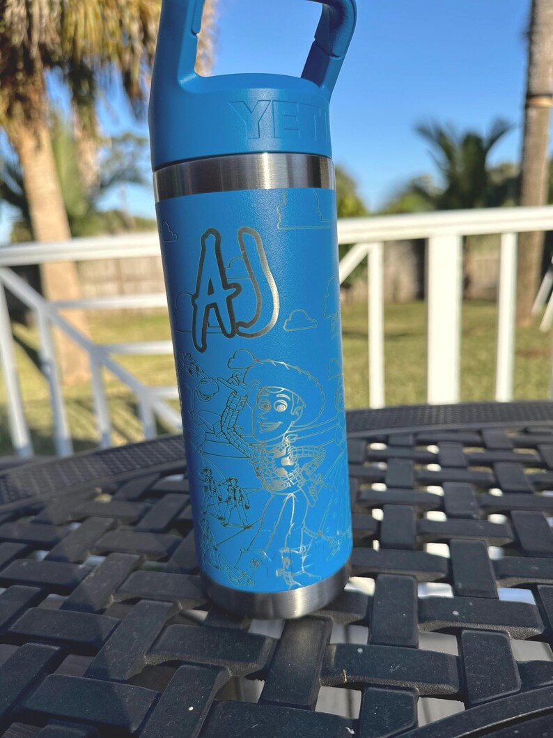 May include: A blue Yeti water bottle with a silver lid and a silver base. The bottle has a white inscription that says "YETI" and a silver inscription that says "AJ". The bottle also has a silver engraved image of Woody from Toy Story.