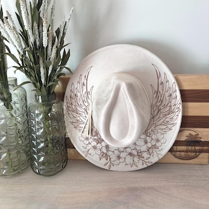 May include: A cream-colored fedora-style hat with a wide brim. The hat features a detailed brown floral and wing design. A decorative tassel hangs from the hat. The hat is resting on a wooden surface.