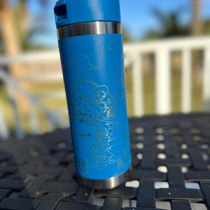May include: A blue stainless steel water bottle with a silver lid and a handle. The bottle has a gold-colored design of characters from the movie Toy Story. The design includes the words "Infinity and Beyond".