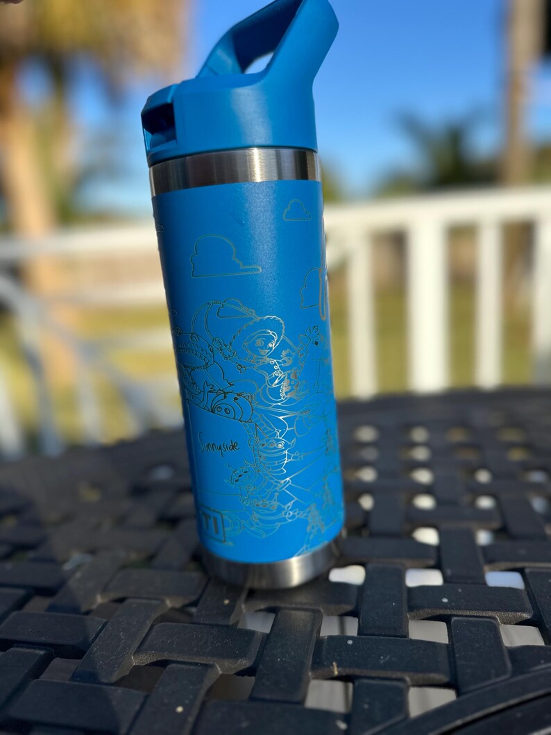 May include: A blue stainless steel water bottle with a silver lid and a handle. The bottle has a gold-colored design of characters from the movie Toy Story. The design includes the words "Infinity and Beyond".