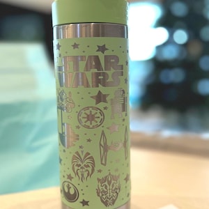 May include: A green stainless steel water bottle with a carrying handle. The bottle is decorated with a silver metallic design featuring the words "Star Wars" and various characters and symbols from the Star Wars franchise.