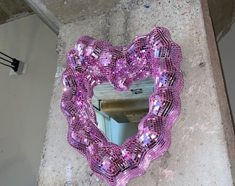 Pode incluir: Um espelho rosa em forma de coração feito de pequenas telhas iridescentes. O espelho está pendurado em uma parede de concreto.