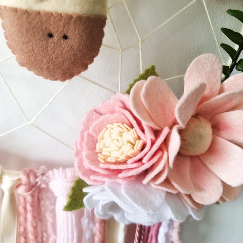 Pode incluir: Uma guirlanda de flores de feltro rosa e branco com folhas verdes. As flores est&atilde;o dispostas em um padr&atilde;o circular e est&atilde;o presas a um cord&atilde;o branco.