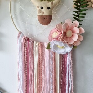 Pode incluir: Um apanhador de sonhos rosa e branco com uma cabe&ccedil;a de girafa de feltro e detalhes florais. O apanhador de sonhos tem um aro de madeira branco e fios de l&atilde; rosa, branco e creme.
