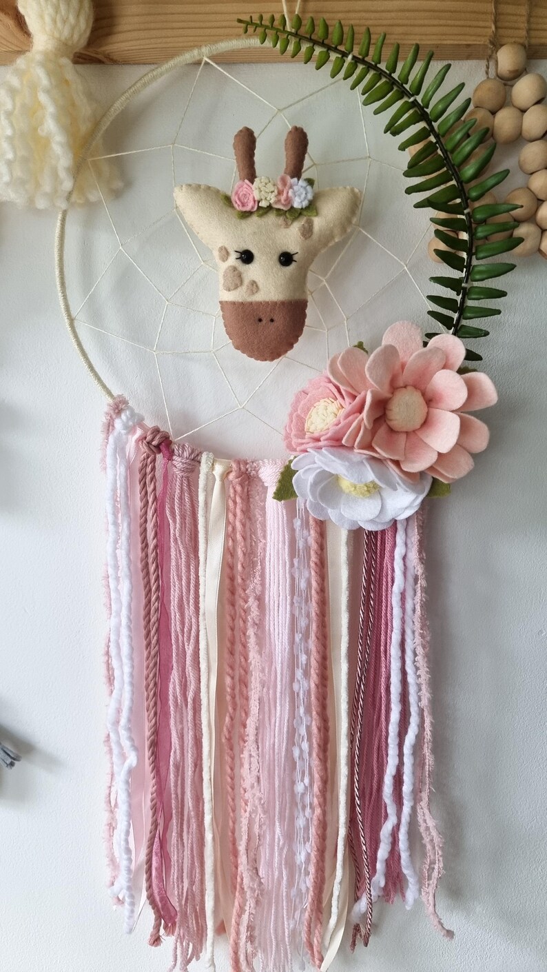 Pode incluir: Um apanhador de sonhos rosa e branco com uma cabe&ccedil;a de girafa de feltro e detalhes florais. O apanhador de sonhos tem um aro de madeira branco e fios de l&atilde; rosa, branco e creme.