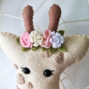 Pode incluir: Uma cabe&ccedil;a de girafa de feltro com chifres marrons e uma coroa de flores. A coroa &eacute; feita de flores de feltro rosa, branco e creme.
