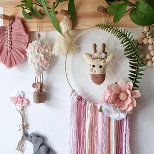 Pode incluir: Um conjunto de decora&ccedil;&atilde;o de ber&ccedil;&aacute;rio tem&aacute;tico floral rosa e branco com um apanhador de sonhos de girafa, um bal&atilde;o de ar quente, um macaco, um elefante e uma parede de penas. O apanhador de sonhos tem borlas de fio rosa e branco. O texto "Mais itens da cole&ccedil;&atilde;o Floral Safari podem ser encontrados na minha loja" est&aacute; na parte inferior da imagem.
