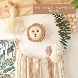 Pode incluir: Um apanhador de sonhos branco com uma cabe&ccedil;a de le&atilde;o de feltro marrom, borlas de fio branco e marrom e pompons brancos. O apanhador de sonhos est&aacute; pendurado em uma vareta de madeira. O texto "Find more Safari nursery decor in my shop" &eacute; vis&iacute;vel na imagem.