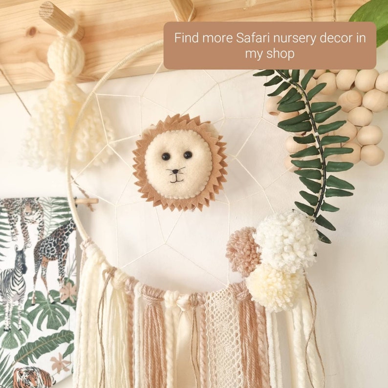 Pode incluir: Um apanhador de sonhos branco com uma cabe&ccedil;a de le&atilde;o de feltro marrom, borlas de fio branco e marrom e pompons brancos. O apanhador de sonhos est&aacute; pendurado em uma vareta de madeira. O texto "Find more Safari nursery decor in my shop" &eacute; vis&iacute;vel na imagem.