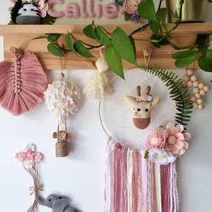 Pode incluir: Um apanhador de sonhos de feltro rosa e branco com um rosto de girafa no centro, rodeado de flores rosa e brancas. O apanhador de sonhos est&aacute; pendurado em uma prateleira de madeira com outras decora&ccedil;&otilde;es de feltro, incluindo um bal&atilde;o de ar quente, um macaco, um elefante e uma zebra.