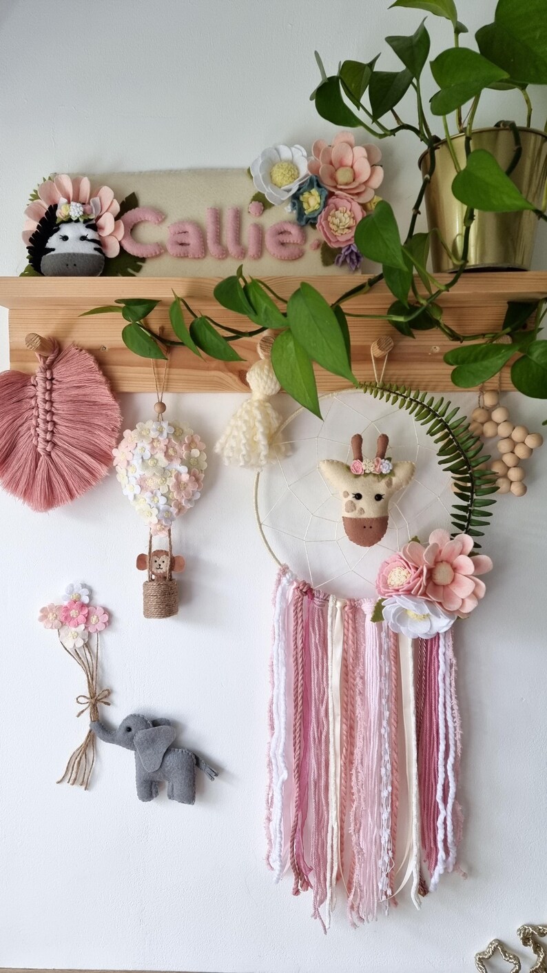 Pode incluir: Um apanhador de sonhos de feltro rosa e branco com um rosto de girafa no centro, rodeado de flores rosa e brancas. O apanhador de sonhos est&aacute; pendurado em uma prateleira de madeira com outras decora&ccedil;&otilde;es de feltro, incluindo um bal&atilde;o de ar quente, um macaco, um elefante e uma zebra.