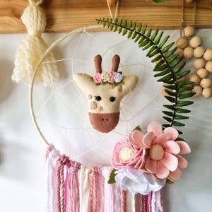 Pode incluir: Um apanhador de sonhos branco com uma cabe&ccedil;a de girafa de feltro, flores rosa e brancas e borlas de fio rosa e branco.