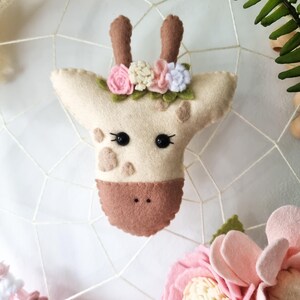 Pode incluir: Uma cabe&ccedil;a de girafa de feltro com um nariz castanho, olhos pretos e uma coroa de flores de feltro rosa, branco e verde.