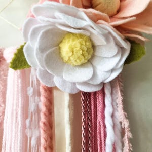 Pode incluir: Uma flor de feltro branco com um centro de feltro amarelo, cercada por flores de feltro rosa e branco. As flores est&atilde;o presas a um fio de fio rosa, branco e creme.