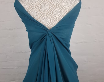May include: Teal fabric draped over a mannequin, showcasing a flowing design. The fabric is gathered and knotted at the center, creating a textured effect. The mannequin's back is visible, with a white lace pattern. The background is a white brick wall.
