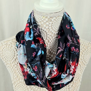 May include: A black infinity scarf with a floral print featuring red, blue and white flowers. The scarf is made of a soft, lightweight fabric.
