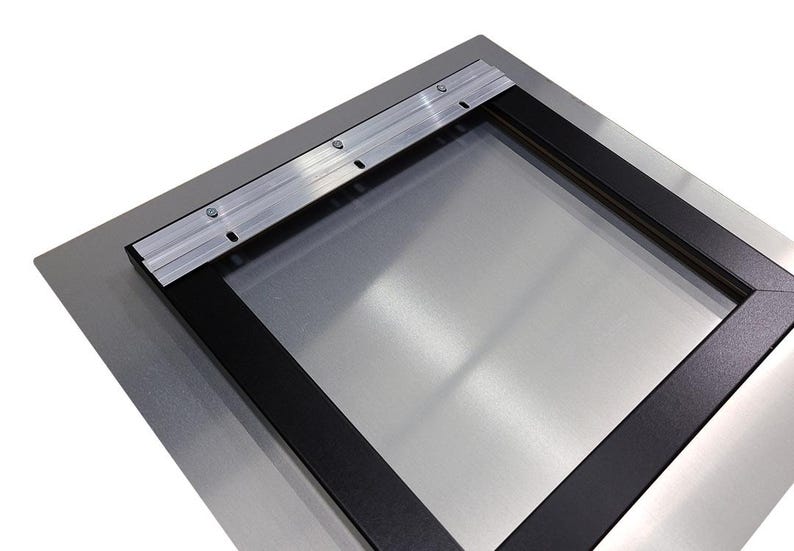 May include: A metal print with a black frame and a silver surface. The back has a metal hanging bracket attached with screws. The frame is rectangular.
