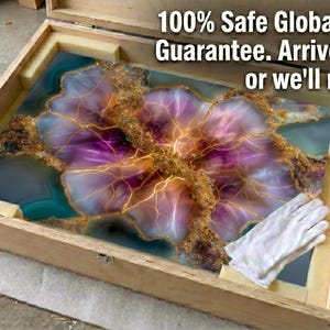 Pode incluir: Uma caixa de madeira retangular contendo uma grande pe&ccedil;a de arte plana e abstrata com padr&otilde;es de resina em tons de roxo, azul-petr&oacute;leo e dourado. O texto 100% Safe Global Delivery Guarantee. Arrive flawless or we'll replace it. est&aacute; impresso.