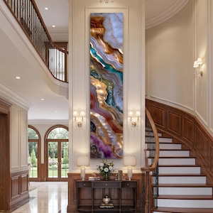 May include: A tall, vertical rectangular wall art piece featuring an abstract agate-inspired design with swirling layers of brown, white, teal, gold, and purple tones, displayed on a central pillar between two staircases in a grand foyer.
