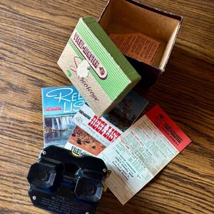 May include: Vintage View-Master stereoscope set with box, reels, and printed materials. The black stereoscope is next to a green and tan box with the words "View-Master Stereoscope". The set includes reel lists and story reels.