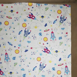 Può includere: Tessuto bianco con un motivo ripetuto di razzi, stelle, pianeti e soli in tonalità di blu, rosso, giallo e verde.