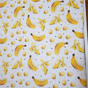 May include: White fabric with a repeating pattern of yellow bananas and small yellow circles. The bananas are in various positions, some are whole and some are peeled.