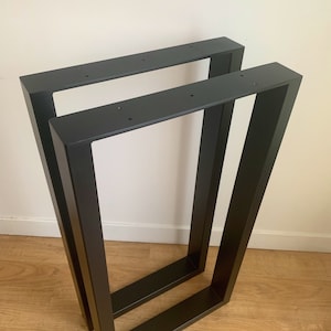 May include: Two black metal table legs with a modern, geometric design. Each leg is rectangular with a trapezoidal shape, featuring pre-drilled holes for easy attachment. The legs are sturdy and suitable for various table sizes.