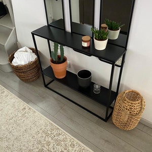 May include: A black metal console table with two shelves, holding potted plants, a candle, and decorative items. A woven basket and a decorative lantern are on the floor. A large mirror is behind the table.