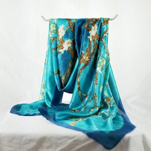 Op de afbeelding: Een turquoise blauwe zijden sjaal met een bedrukt ontwerp van een bloeiende amandelboomtak met witte bloemen. De sjaal is over een zilveren hanger gehangen.