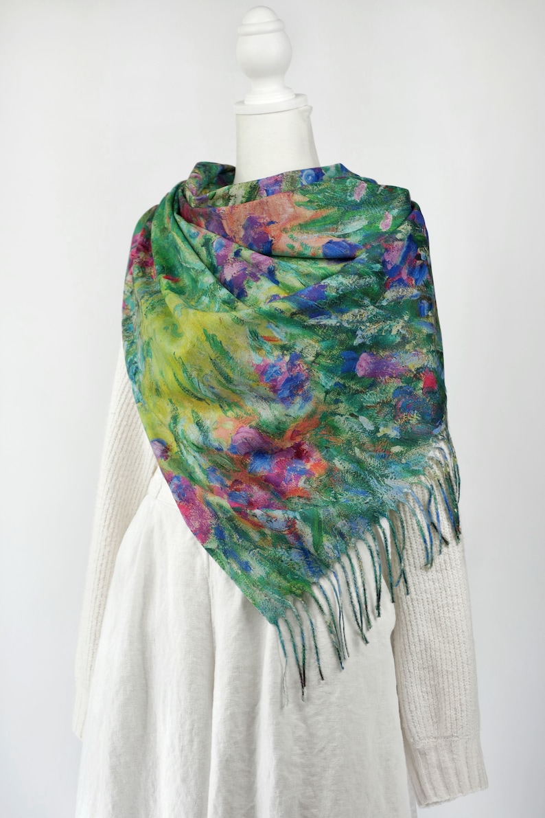 May include: A colorful scarf with a floral design in shades of green, blue, pink, and yellow. The scarf has a fringed edge and is draped over a white sweater and dress form.
