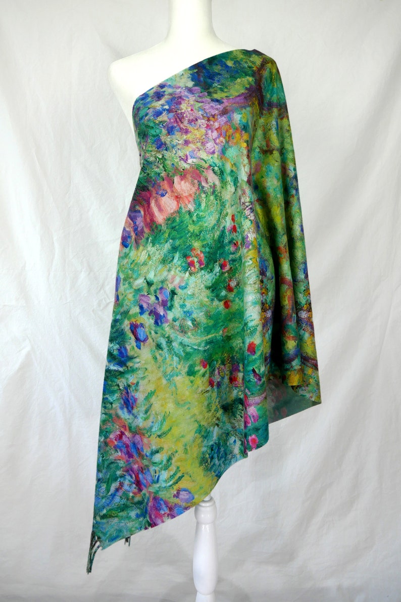 May include: A one-shoulder garment draped over a mannequin, showcasing a vibrant floral print. The fabric features a lush garden scene with green, yellow, pink, and purple hues, reminiscent of an impressionist painting. The garment is likely a scarf or shawl.