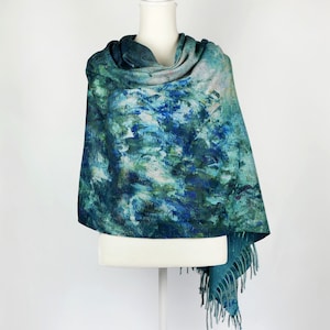 K&ouml;nnte beinhalten: Ein t&uuml;rkis-blauer Schal mit einem malerischen Design. Der Schal ist &uuml;ber einer wei&szlig;en Schaufensterpuppe drapiert und zeigt das abstrakte Muster aus Blau, Gr&uuml;n und Wei&szlig;. Der Schal hat einen Fransenrand. Das Gesamtthema ist k&uuml;nstlerisch und tragbar.