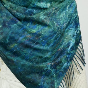K&ouml;nnte beinhalten: Ein t&uuml;rkis-blauer Schal mit malerischem Design. Der Schal hat einen Fransenrand und ist &uuml;ber einem wei&szlig;en Pullover drapiert. Das Muster des Schals &auml;hnelt einer abstrakten Landschaft mit Pinselstrichen in verschiedenen Blau- und Gr&uuml;nt&ouml;nen.