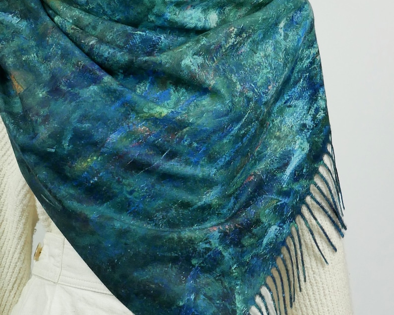 K&ouml;nnte beinhalten: Ein t&uuml;rkis-blauer Schal mit malerischem Design. Der Schal hat einen Fransenrand und ist &uuml;ber einem wei&szlig;en Pullover drapiert. Das Muster des Schals &auml;hnelt einer abstrakten Landschaft mit Pinselstrichen in verschiedenen Blau- und Gr&uuml;nt&ouml;nen.