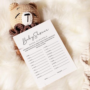 Pode incluir: Um cart&atilde;o branco com o texto "Baby Shower Word Scramble" impresso em preto. O cart&atilde;o tem uma lista de palavras embaralhadas relacionadas a beb&ecirc;s. O cart&atilde;o est&aacute; sobre uma superf&iacute;cie branca e fofa com um urso de pel&uacute;cia de croch&ecirc; e um anel de madeira.