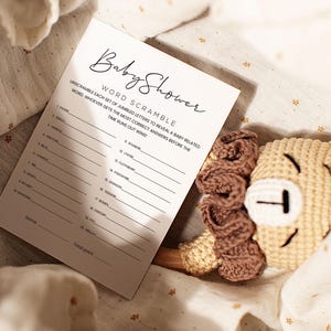 Pode incluir: Um cart&atilde;o branco com o texto "Baby Shower Word Scramble" e instru&ccedil;&otilde;es para decifrar palavras relacionadas com beb&eacute;s. O cart&atilde;o est&aacute; sobre um tecido bege com um brinquedo de le&atilde;o de crochet.