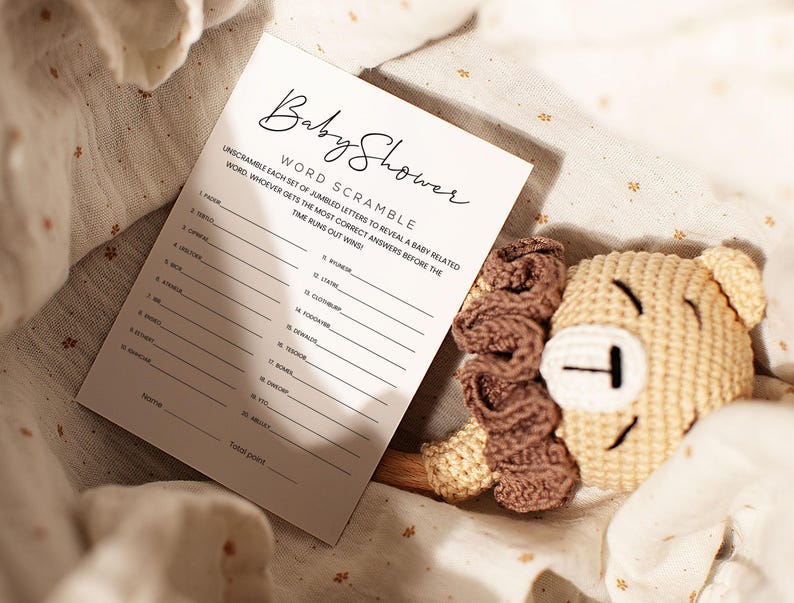 Pode incluir: Um cart&atilde;o branco com o texto "Baby Shower Word Scramble" e instru&ccedil;&otilde;es para decifrar palavras relacionadas com beb&eacute;s. O cart&atilde;o est&aacute; sobre um tecido bege com um brinquedo de le&atilde;o de crochet.