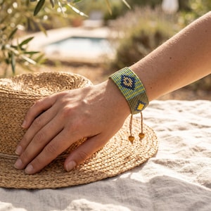 Puede incluir: Una pulsera tejida con un patrón geométrico en tonos verde oliva, azul claro y amarillo. La pulsera tiene dos cordones colgantes terminados con pequeñas cuentas de madera en los extremos, lo que permite un ajuste regulable en la muñeca.