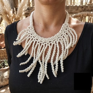 Può includere: Collana in macramè color crema con un design intrecciato e frange. La collana è realizzata in spessa e morbida corda di cotone e viene indossata su un top nero. La collana ha uno stile bohémien.