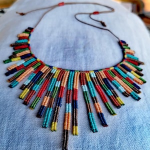 Può includere: Una colorata collana in macramè con un design a pettorina. La collana presenta una varietà di colori, tra cui rosso, blu, arancione e verde. È realizzata con piccole perline fittamente intrecciate e ha un cordino marrone per regolare la lunghezza.